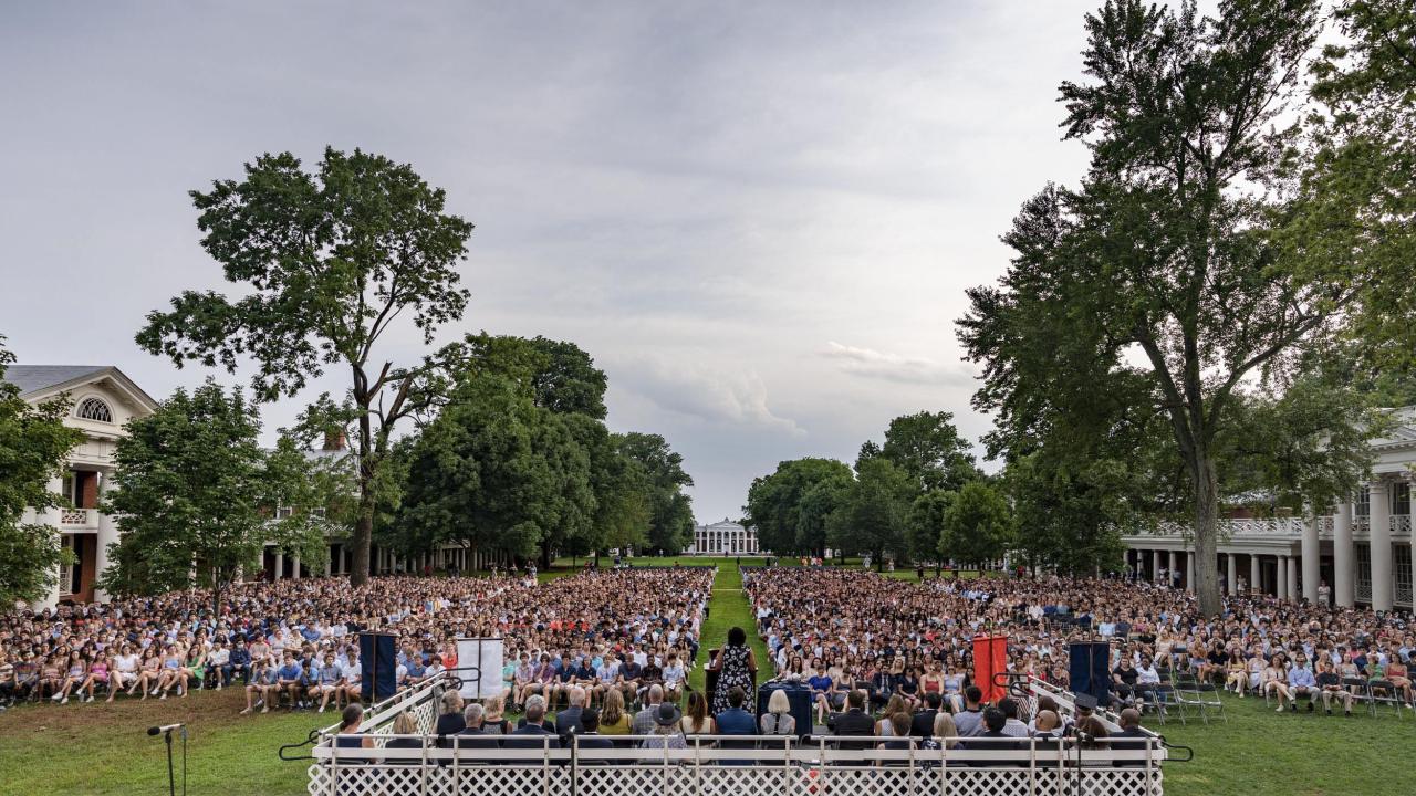 Students sitting in chairs on the lawn looking at a stage where a woman is giving a speech